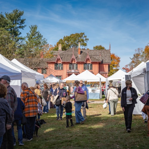 People wander through a market with white tents. A Victorian Gothic-style house with lush trees is seen in the background.