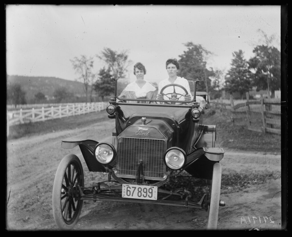 Two people in an early 20th-century Ford Model T car on a rural dirt road, flanked by white fences and lush trees.