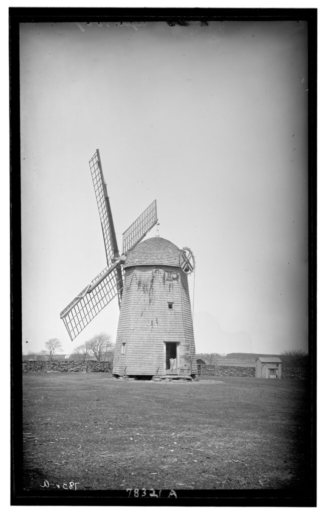 Historic wooden windmill with large sails, stands solitary on a field. Sparse trees and stone walls in the background.
