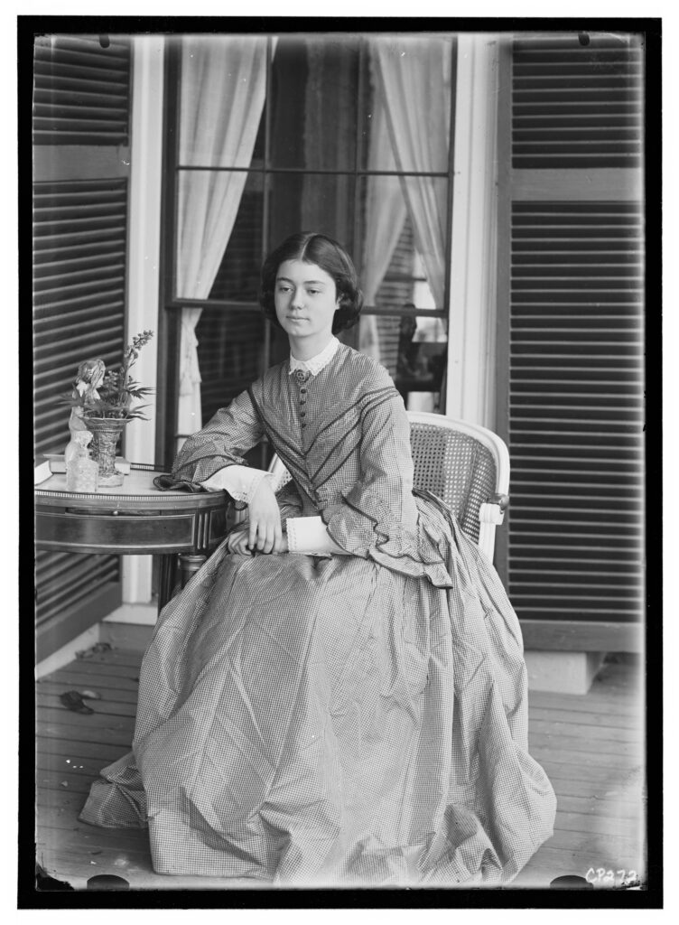 Seated woman in a Victorian dress on a porch with shutters, by a table with a lush plant and lace curtains behind.
