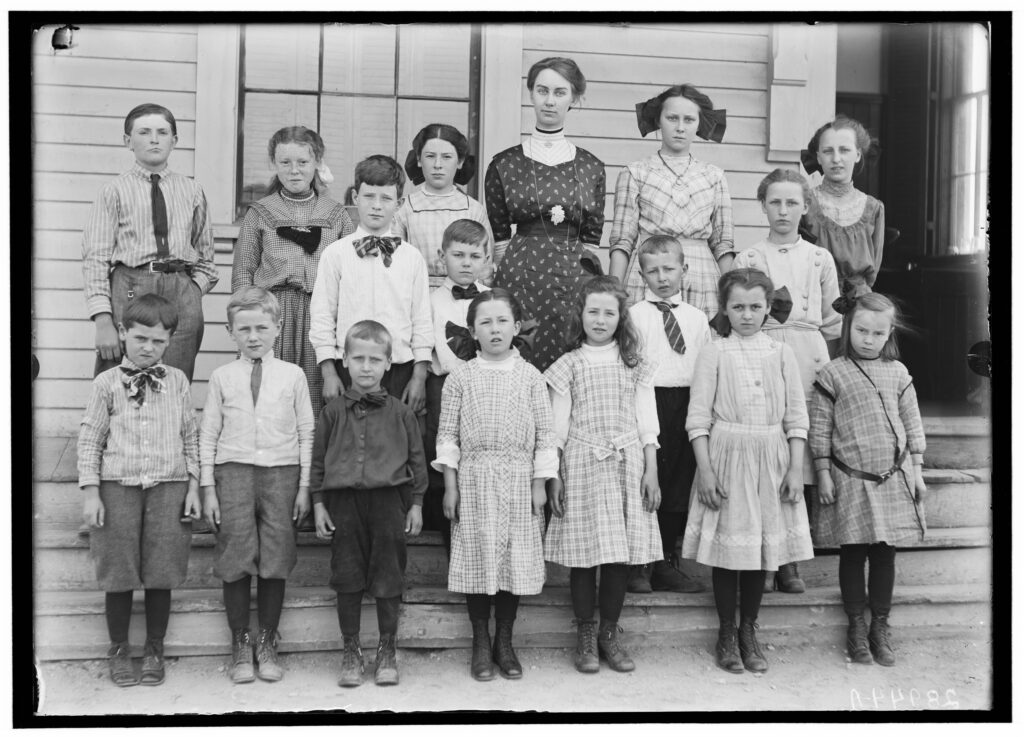 Historic photo of a teacher with her class on wooden steps, wearing period clothing in early 20th-century architectural setting.