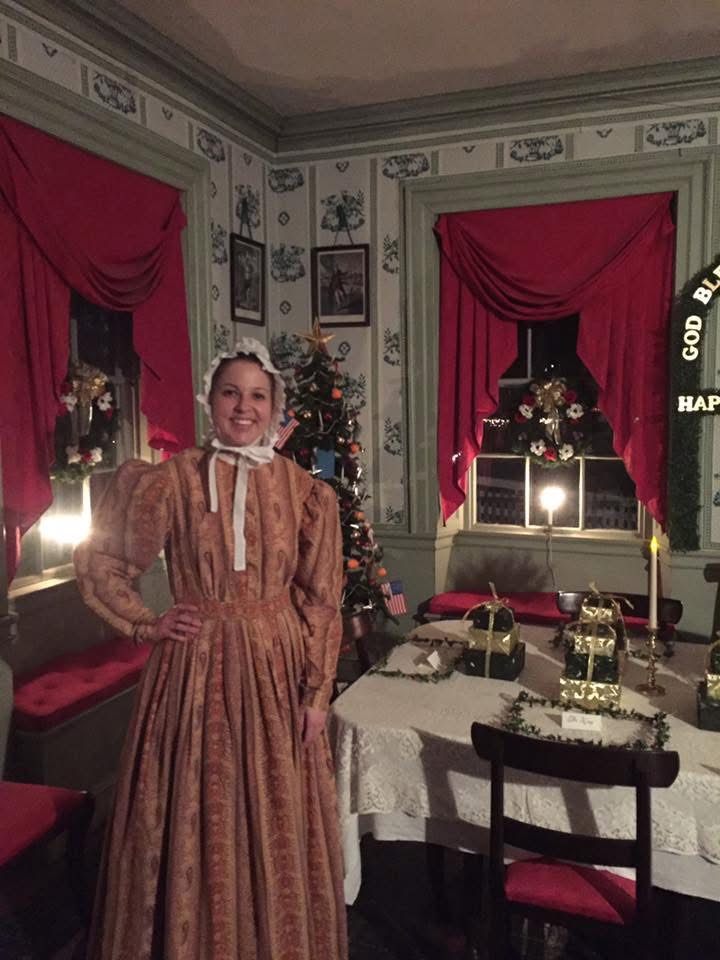 A person in a vintage dress stands by a table in a historic room with red drapes, wallpaper, and Christmas decorations.