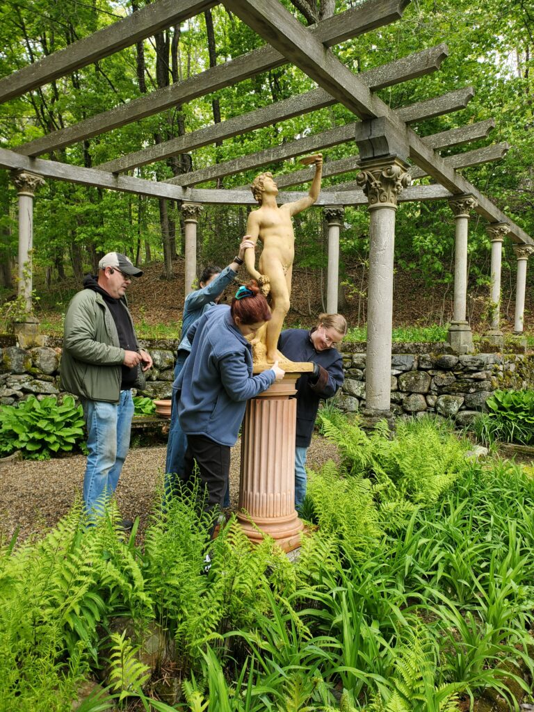 People examine a classical statue under a pergola, surrounded by lush greenery.