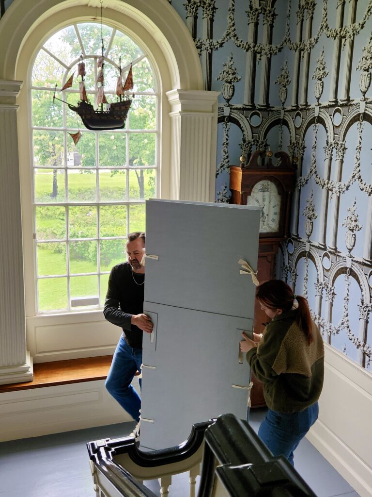 Two people move a large panel up a staircase in a room with ornate wallpaper and a grand arched window featuring a ship model.