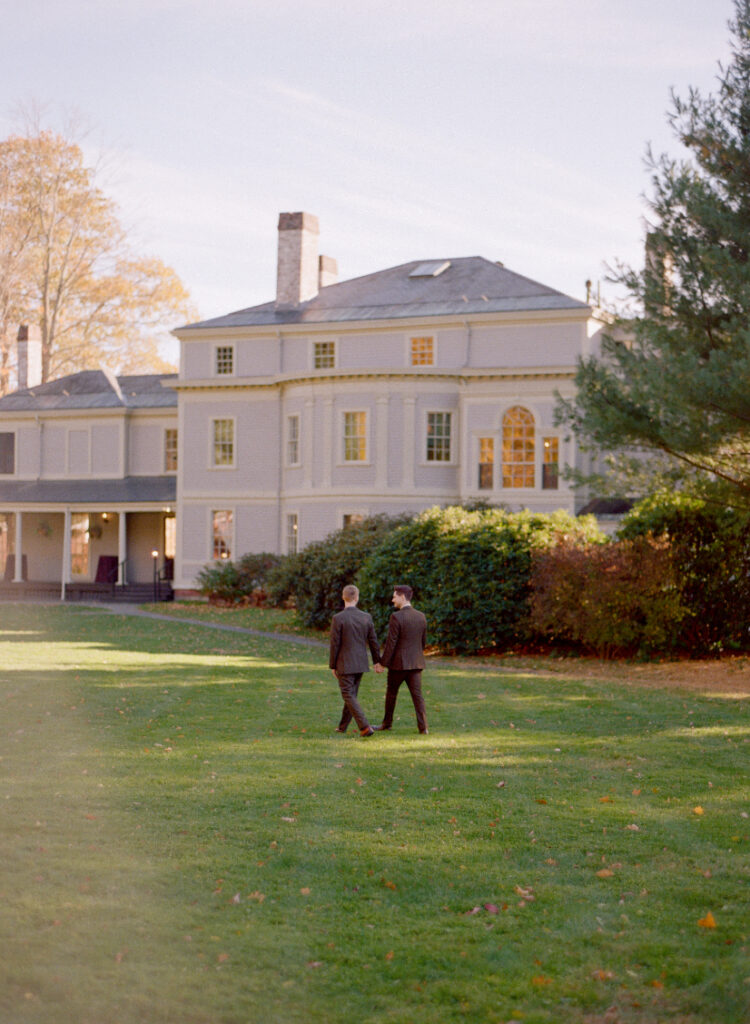 Georgian-style house with tall windows, chimneys, and lush surroundings. Two people walk on the green lawn.