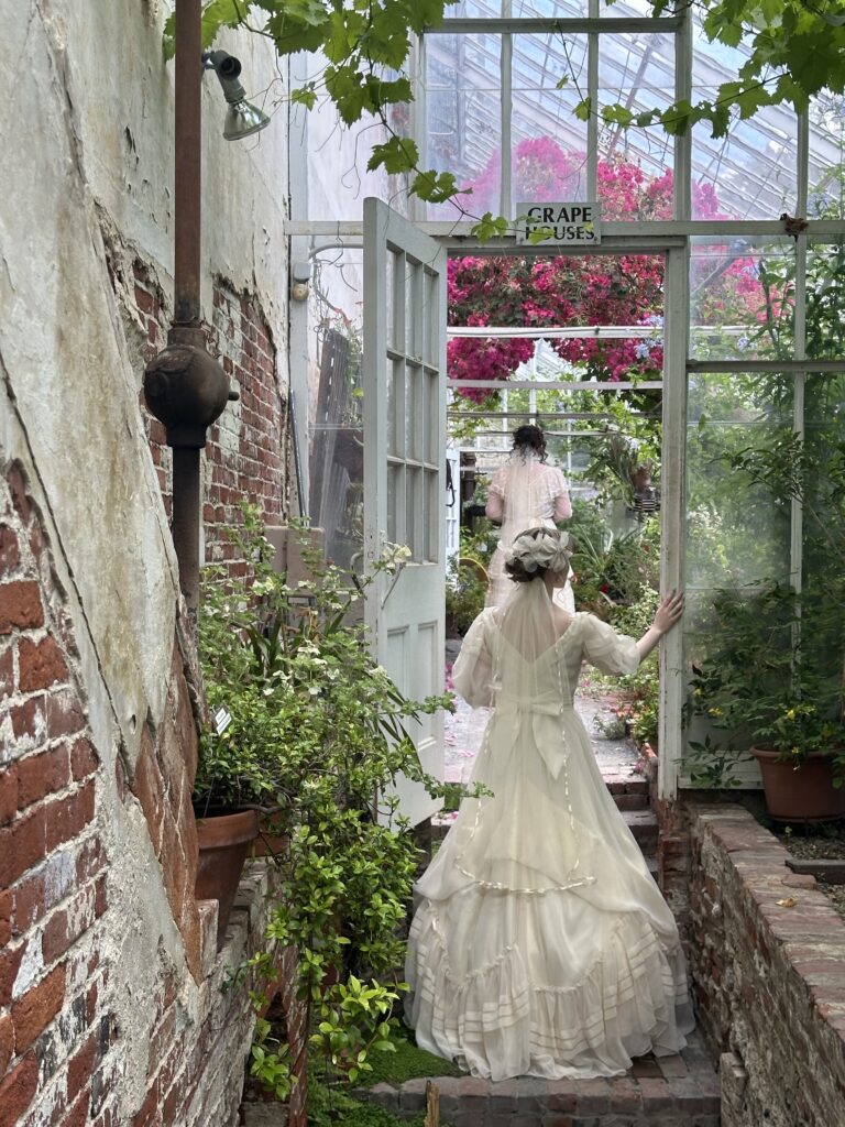Victorian-dressed woman explores a rustic, brick-lined greenhouse with lush greenery and bright flowers beyond a glass door.