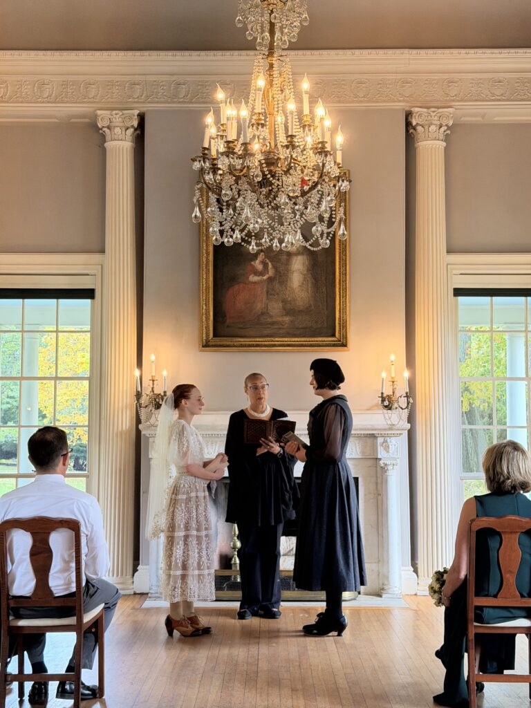A wedding ceremony in an elegant room with neoclassical columns, a grand chandelier, and an ornate fireplace.