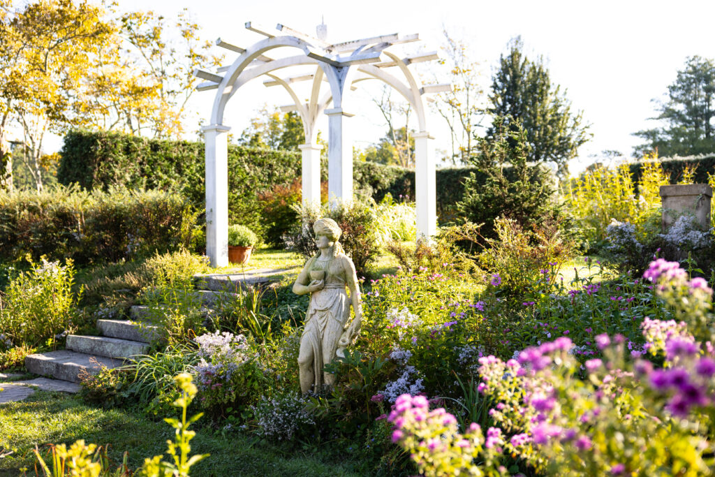A classical statue stands in a lush garden, framed by a white pergola and surrounded by vibrant flowers and greenery.