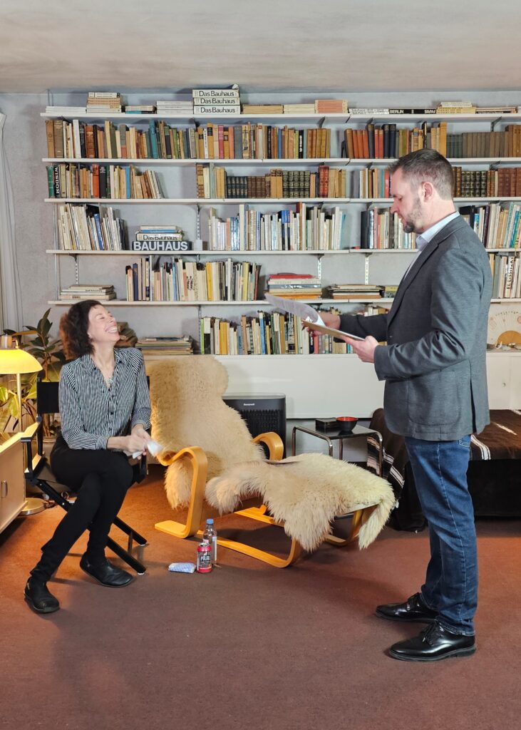Two people converse in a library, surrounded by books on Bauhaus design. A sheepskin-covered chair is in the foreground.