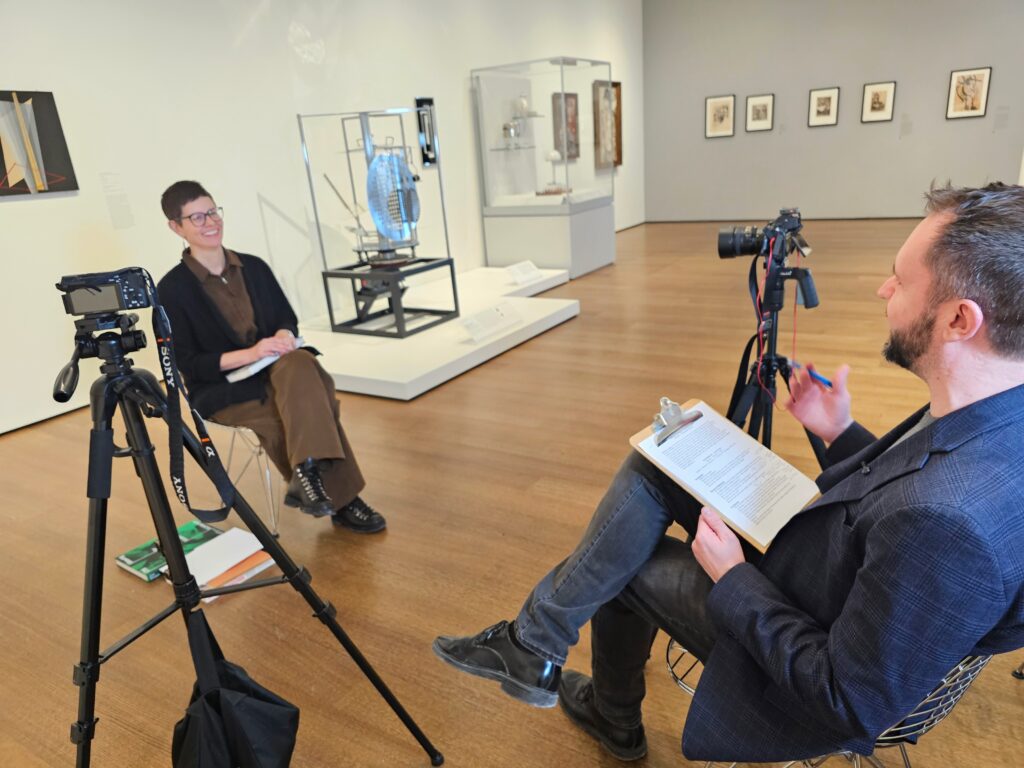 Two people engage in an interview in a modern art gallery, surrounded by framed artwork and sculptures.
