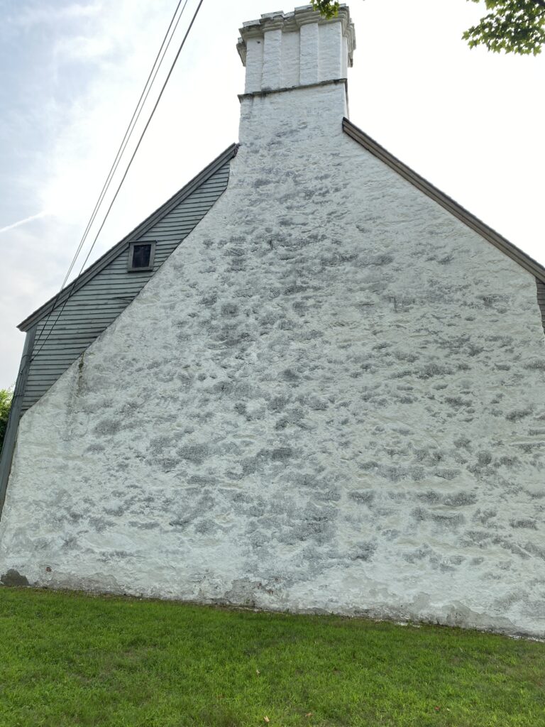 Historic stone chimney against weathered wood siding, surrounded by lush grass, stands under a clear sky.