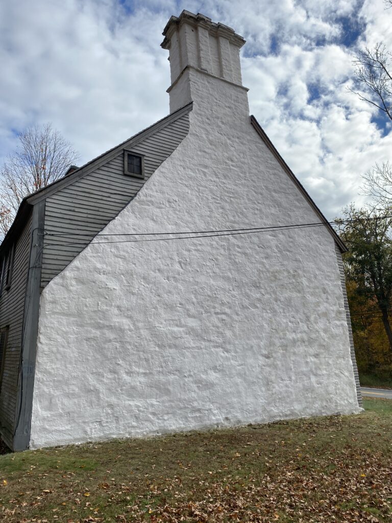 Historic house with a distinctive white stuccoed chimney, contrasting weathered gray siding, surrounded by autumn trees.