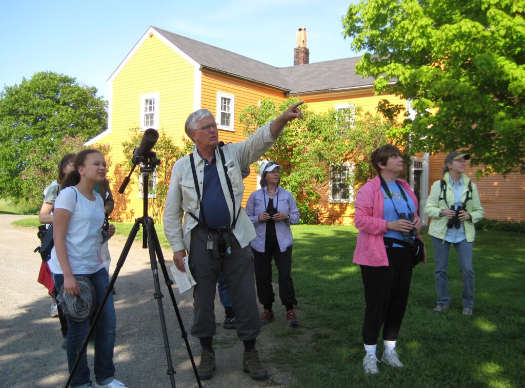A group studies nature outside a bright yellow farmhouse, guided with binoculars and telescopes. Lush greenery surrounds them.