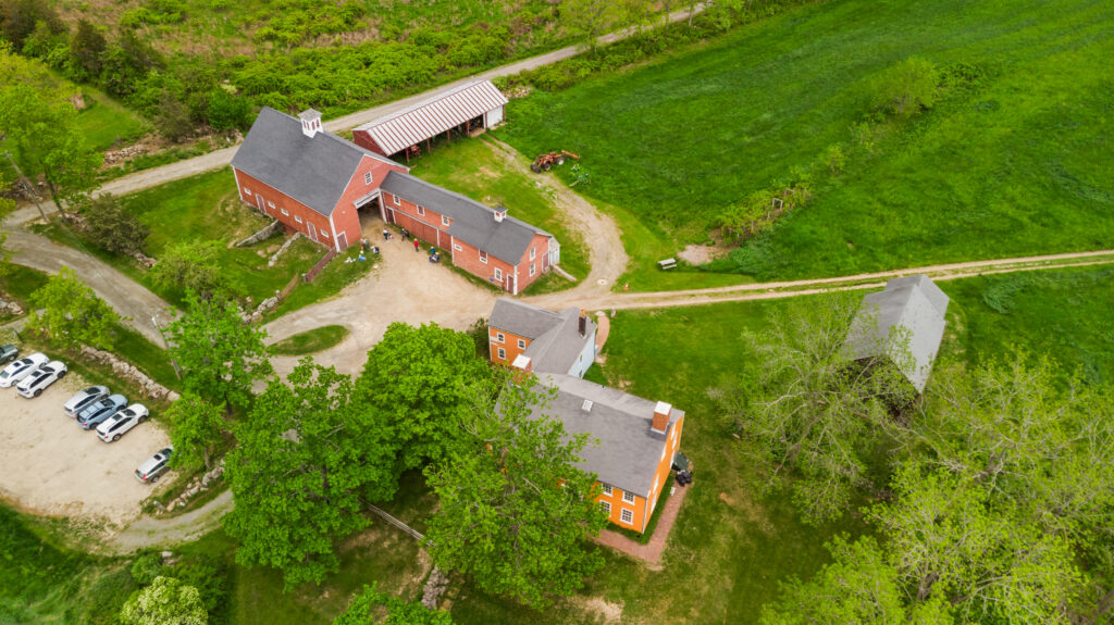 Aerial view of historic red barns and a house amidst lush greenery, with cars parked on a gravel path.