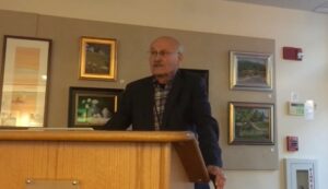 Man speaking at podium in art gallery, surrounded by framed paintings on the wall.