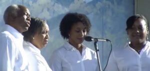 Four people singing in a choir, dressed in white shirts, standing in front of a microphone with a blue sky backdrop.
