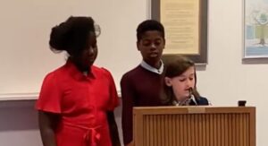 Three children give a speech at a podium in an institutional setting.