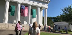 People gather outside a white church with banners and the American flag under a clear blue sky.