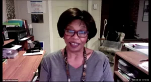 A person with glasses smiling during a video call in a cozy home office setting.