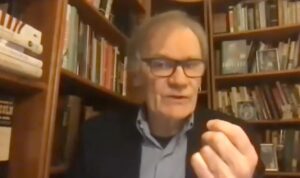 Man speaking in a library setting, gesturing with hand, surrounded by bookshelves.