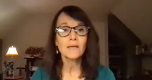 Woman in glasses speaking during an online meeting, warmly lit room with shelves and a lamp in the background.