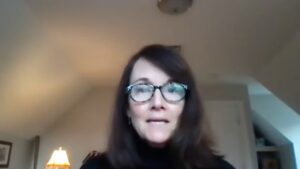 Woman in glasses speaking during a video call, dressed in black, with a lit lamp in the background.