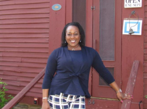 Smiling woman in navy cardigan standing in front of red building with a Open June-Sept sign.