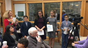 Students presenting in front of a camera and audience at a school event, holding papers and dressed formally.
