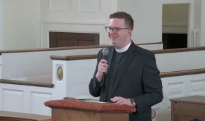 Man in clergy attire speaking at a church podium, holding a microphone, smiling.