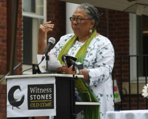 Woman speaking at Witness Stones Old Lyme event, wearing glasses and green scarf, standing at a podium with microphone.