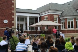 Outdoor speech at historic brick building with audience, cameraman, and speaker under a large umbrella.