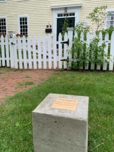 Historic yellow house with white picket fence, brick path, and engraved stone plaque in the foreground.
