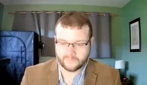 A person with glasses and headphones sits in a home office with green walls and a blue tent in the background.