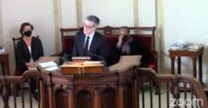 Man speaking at a courtroom podium, two masked women seated nearby, Zoom logo visible.
