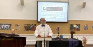 Man giving a lecture on Northern slavery history, titled Witness, in a room with images and a piano.