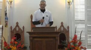 Man speaks at podium in a church setting with colorful floral arrangements and wooden chairs in the background.