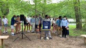 Outdoor school event with students holding certificates, standing in a group near a speaker, surrounded by trees.