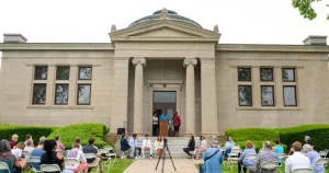Outdoor ceremony at historic building with attendees seated and speakers at podium.