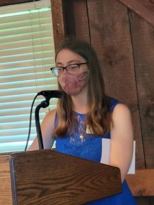Person in blue dress speaking at podium with microphone, wearing a mask, indoors near wooden wall and window blinds.
