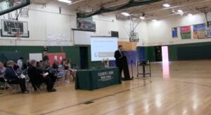 School event in gymnasium with speaker at podium, attendees seated, and presentation screen visible.