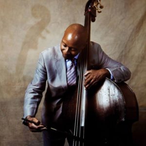 Musician in a suit playing an upright bass with a bow against a neutral background.