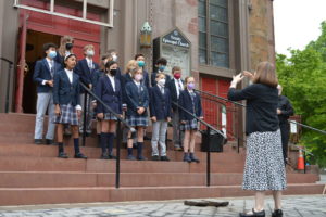 Church choir group with masks singing on steps, director conducting them outside Trinity Episcopal Church.