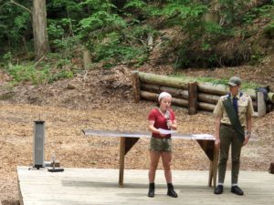 Two people at a wooden podium in a forest setting, one speaking into a microphone, the other in a scout uniform listening.