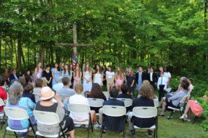Students gathered in an outdoor ceremony with audience seated, surrounded by lush green trees.