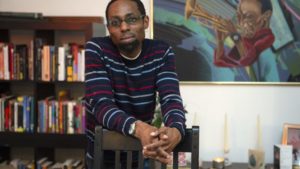 Man in striped sweater leans on chair in cozy room with bookshelves and colorful trumpet painting, creating a relaxed atmosphere.