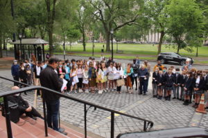 A speaker addresses a diverse group of students gathered outdoors near a park.