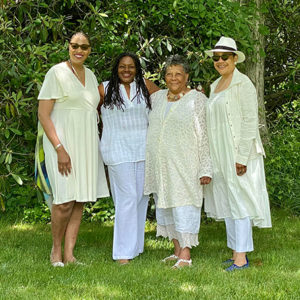 Four women in white outfits smiling in a lush garden setting on a sunny day.