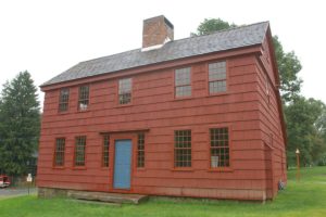 Historic red colonial house with a blue door, surrounded by lush greenery and trees.