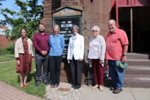 Group outside St. Paul's Episcopal Church, smiling by entrance sign, sunny day.