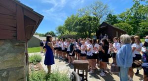 A group of students standing outdoors, attentively listening to a woman in a blue dress.